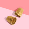 Two gold thimbles with a textured surface placed on a two-tone background, one thimble standing upright and the other lying flat.
